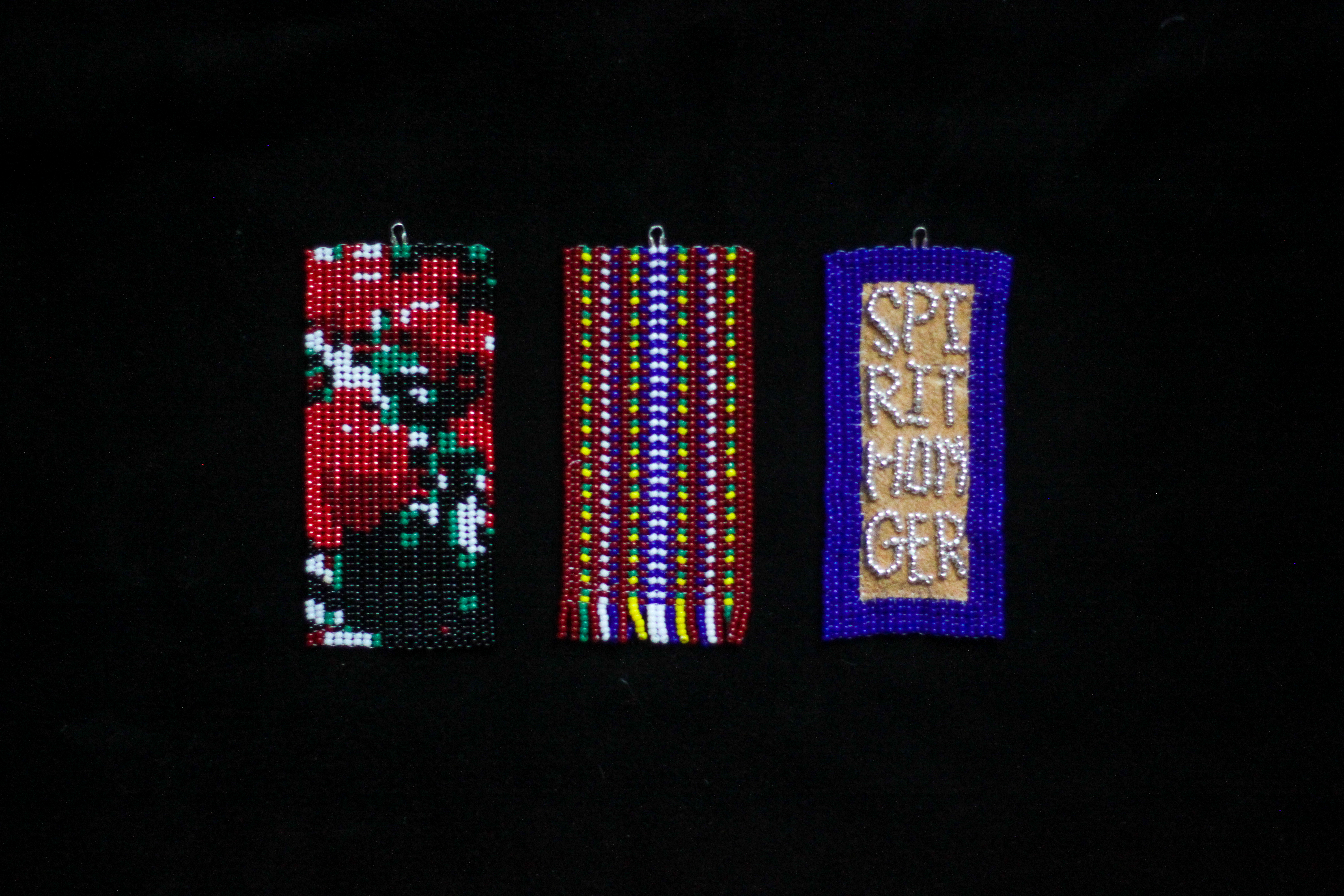 A close-up shot of three rectangular beaded tokens on black hide. The left-most depicts a rose bush; the centre-most depicts a Métis sash; the right-most features the words "spiritmonger" on hide, surrounded by cobalt beads.