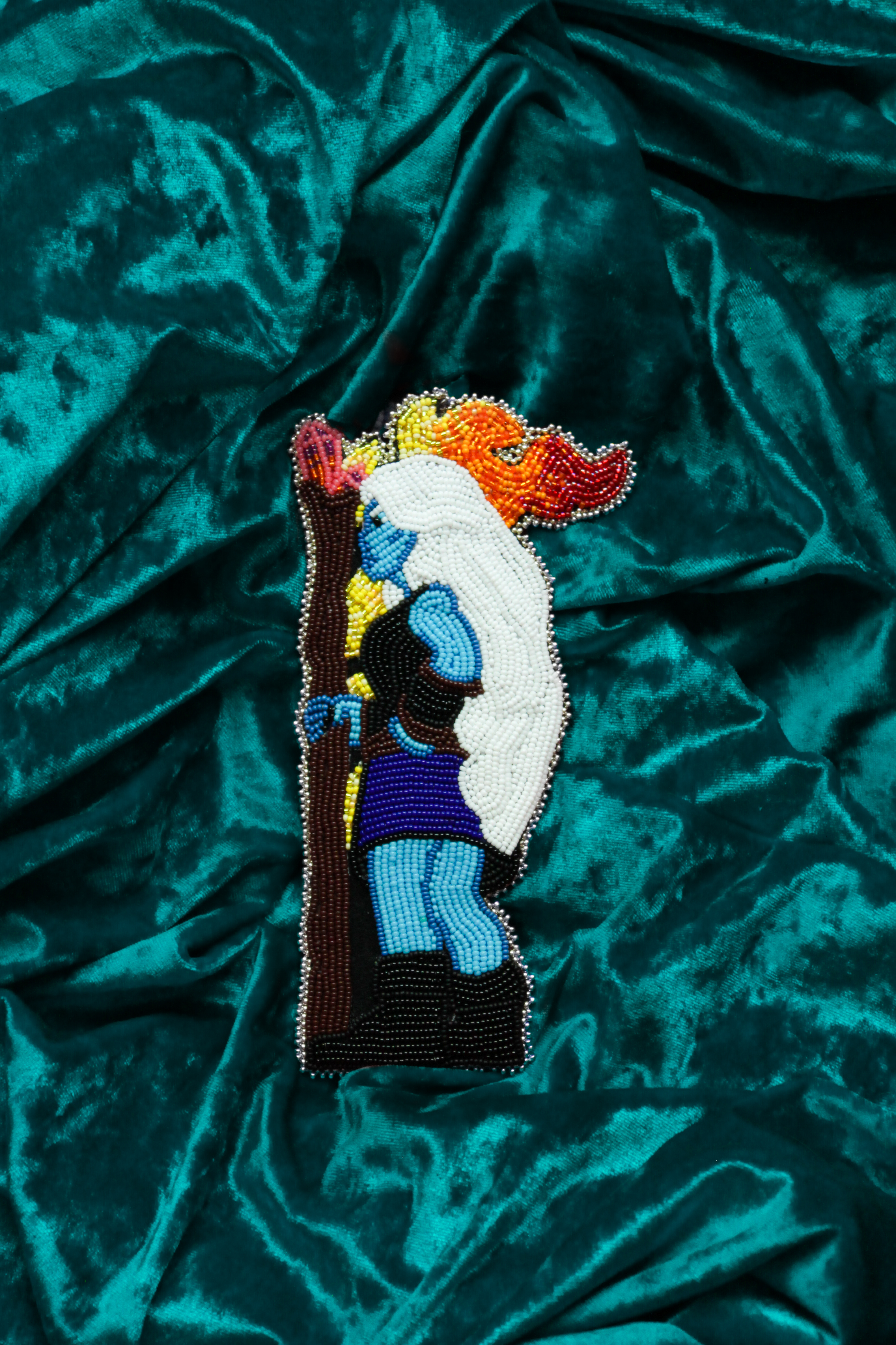 A beaded portrait of a fantasy character with blue skin and long white hair, holding a wooden staff with fire coming from the top. It is resting on a stretch of teal velvet.