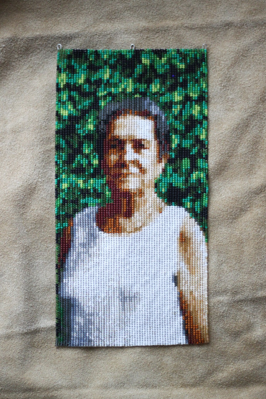 A beaded portrait of a Métis woman with grey hair wearing a white shirt standing in front of a green, leafy hedge. The portrait is resting on tan-coloured hide.
