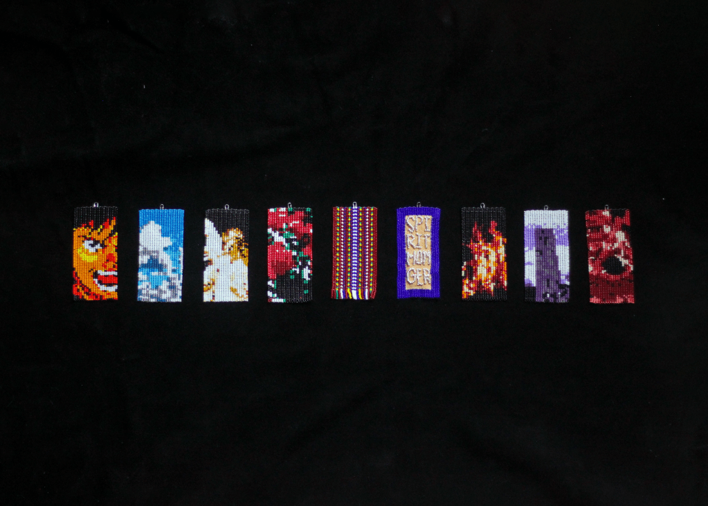 Nine beaded tokens stacked in a single row, depicting faces, a blue sky, an angel, roses, a Métis sash pattern, fire, and a tower. They rest on a stretch of black hide.