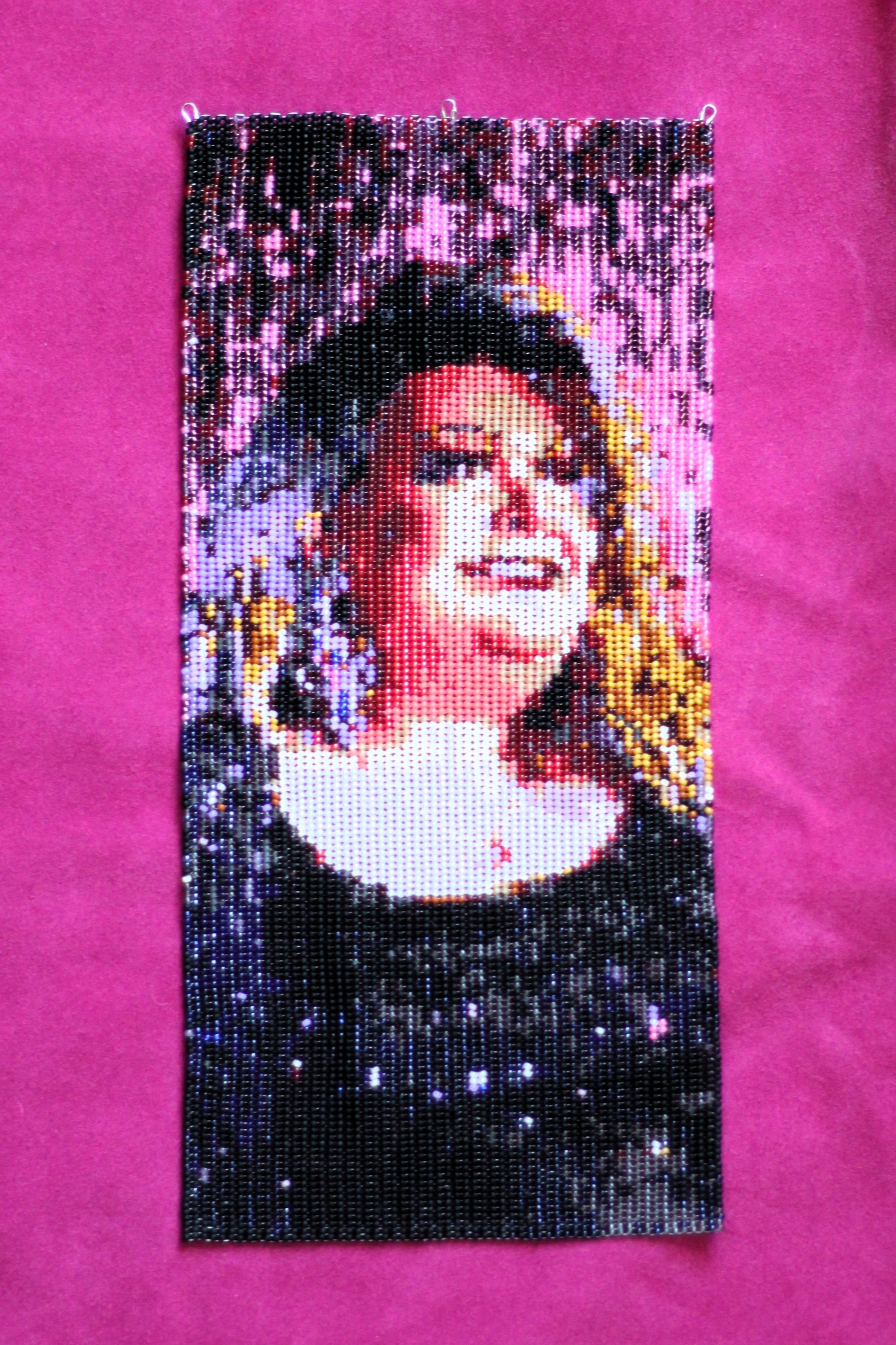 A beaded portrait of a Métis drag queen with blonde hair wearing a sparkly black top in front of a pink backdrop. The portrait is resting on pink hide.