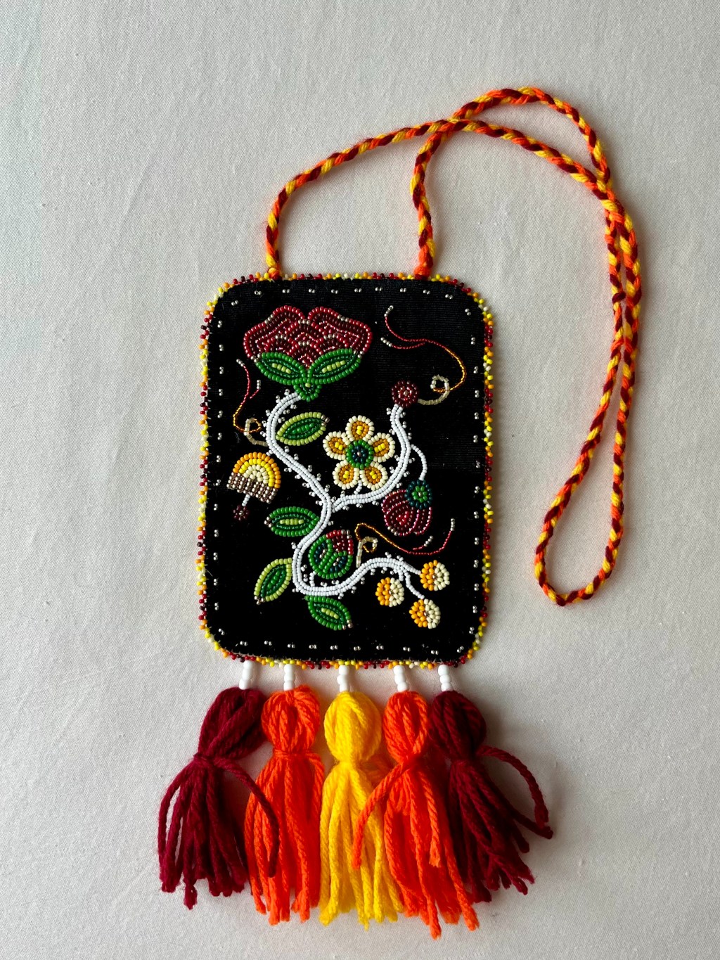 A photo of a medallion. Métis-style floral beadwork is embroidered on black velvet. Five poms made of yarn hang from the bottom; the necklace portion is created by braided yarn.